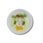 Prato de cerâmica branco, quadrado, com um desenho de um rosto de menina sorrindo, decorado com brócolis como cabelo e outros alimentos para formar um rosto divertido.