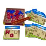 Jogo Baú do Tesouro da Materiais para Brincar, mostrando um baú de madeira aberto com pérolas douradas e peças de letras e números. Uma pinça está sendo usada para pegar uma das peças.