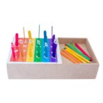 Kit Caixa de Palitos Coloridos da Materiais Para Brincar, mostrando uma caixa de MDF com alguns palitos já inseridos nos orifícios. Uma cartela com um desafio de cores está visível ao lado.