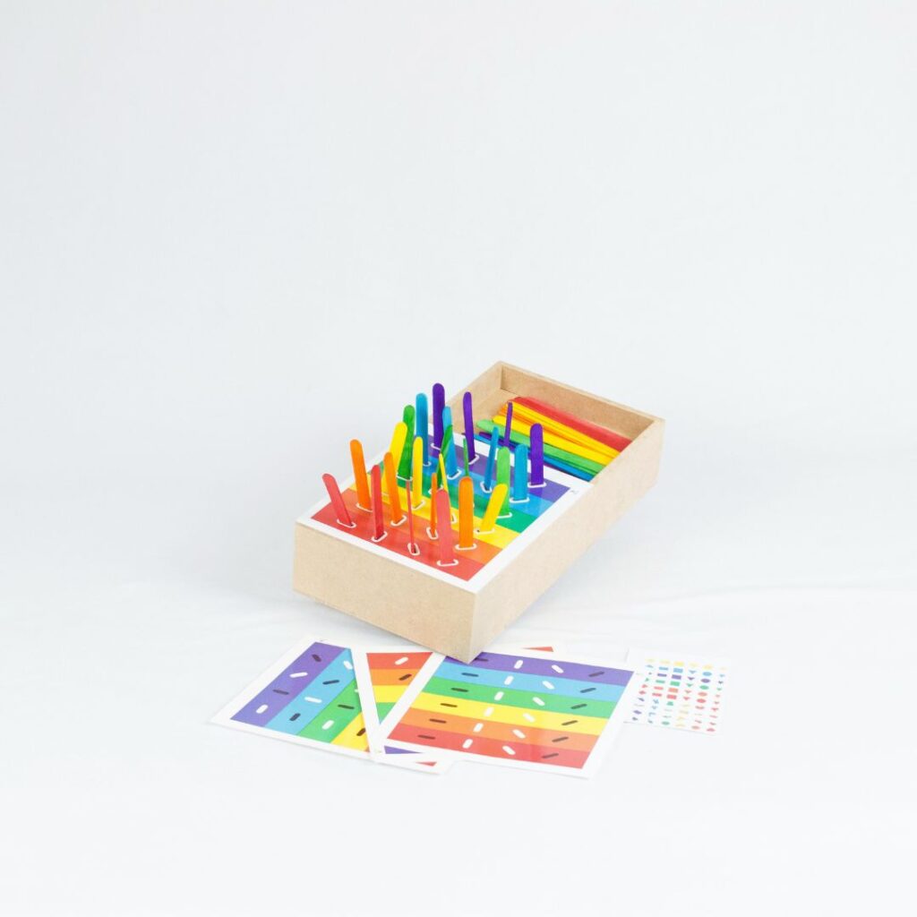 Kit Caixa de Palitos Coloridos da Materiais Para Brincar, mostrando uma caixa de MDF com alguns palitos já inseridos nos orifícios. Uma cartela com um desafio de cores está visível ao lado.