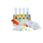 O jogo Sequência de Pompons da Materiais Para Brincar, mostrando a caixa de madeira com os 4 tubos de ensaio. Uma mão de criança está usando a pinça bola para colocar um pompom colorido em um dos tubos, seguindo o padrão de uma das fichas modelo.