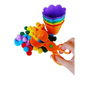 Kit Tesoura, Pompons e Potes Coloridos da Materiais Para Brincar, mostrando uma tesoura de transferência sendo usada por uma mão de criança para pegar um pompom azul de uma tigela e colocá-lo em um pote da mesma cor.